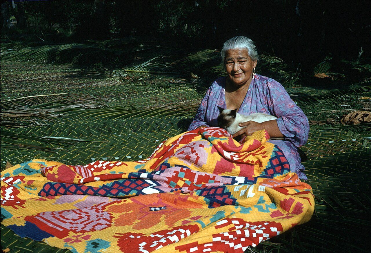 Woman sitting on a kikau (coconut frond mat) making a tivaevae (quilt). She has a siamese cat on her lap. Photograph taken on Rarotonga, Cook Islands.