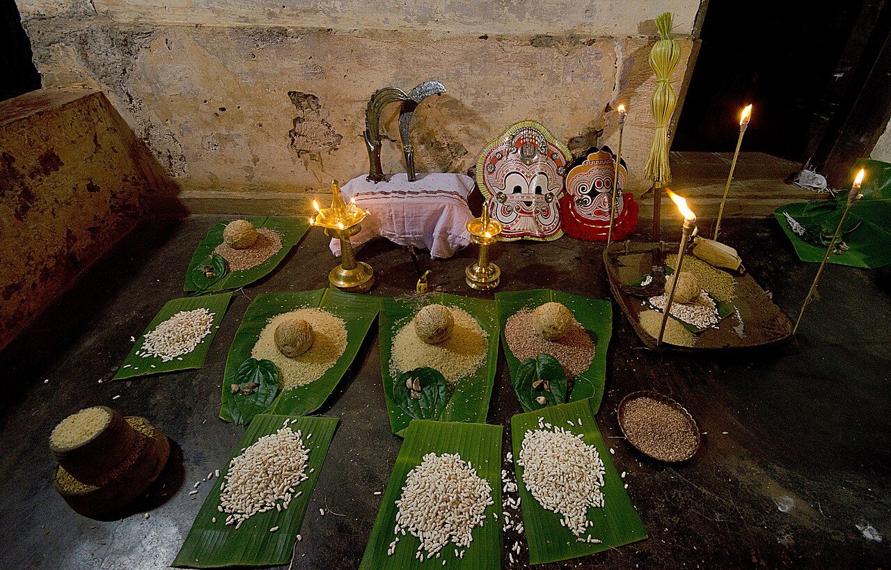 Offerings for Pottan Theyyam.Rice,Coconut,Arcanut etc are kept on plantain leaves . Three Sickles of Pottan Theyyam are also seen. The mask on the left side belongs to Pottan Theyyam and the smaller mask on right side is of Polaran Theyyam.