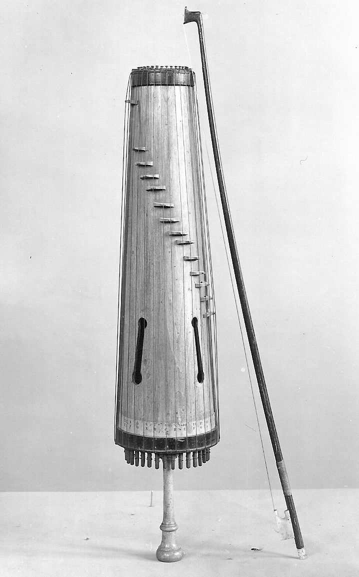 Bowed zither with sixteen wire strings passing over individual moveable wood bridges arranged on a diagonal line over the curved belly; the body trapezoidal in outline and half-circular in cross-section, being a longitudinal half section of a tapering hollow cylinder of pine;…
