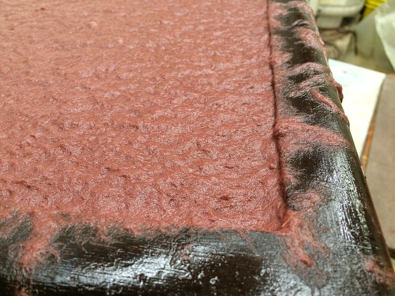 This image shows red pigmented flax fiber on a paper mould, still contained by a deckle. This sheet is quite thick which is why there is fiber spilling onto the deckle.