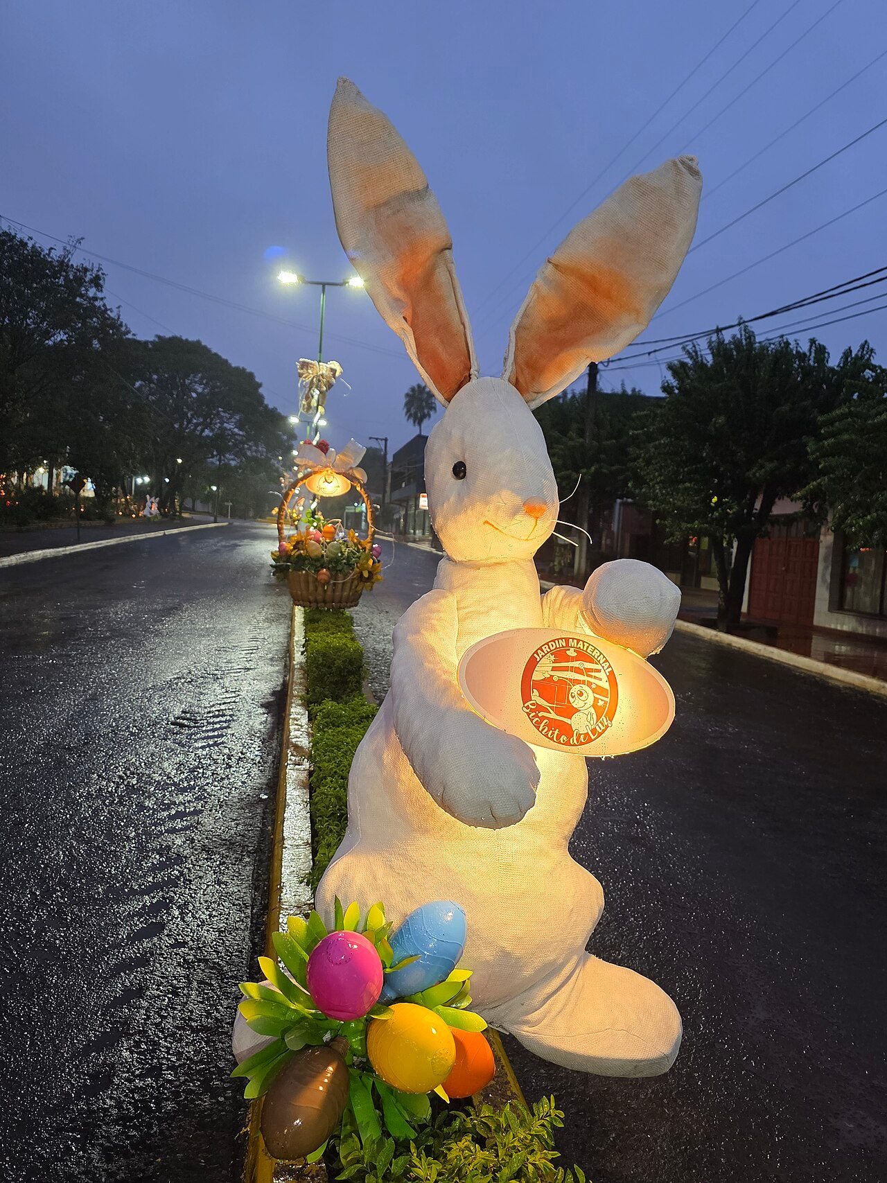 Conjunto de decoraciones y alegorías alusivas a la Pascua 2026 instaladas en la vía pública de Capioví, sobre la avenida San Luis Gonzaga, registradas durante una madrugada con lluvia, donde se aprecian ornamentos temáticos, estructuras escenográficas y ambientación festiva…