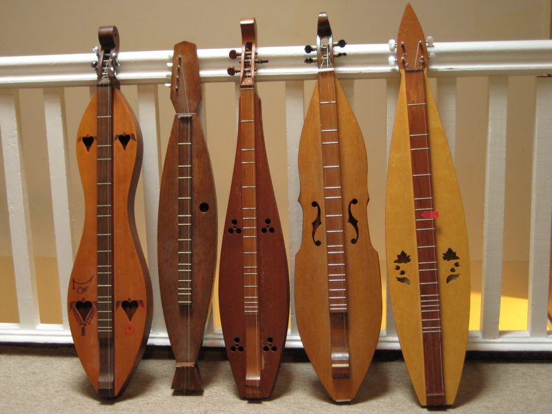 A variety of shaped of Appalachian dulcimer . I don't recall all the makers, but the small dark-brown one is a Hughes, the violin-shaped one is a Pete Wheeler, and the leaf-shaped one is a Cedar Creek. The one on the far left with the painting is out of some small outfit in…