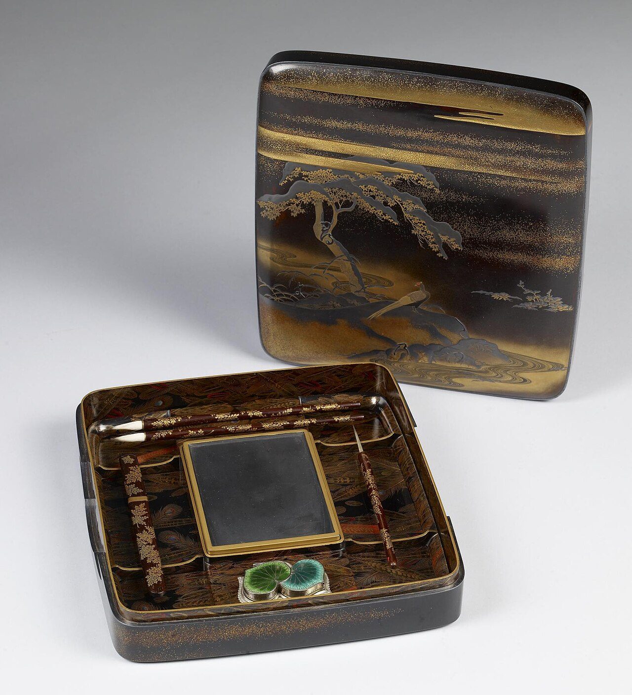 Implements inside the box: A rectangular inkstone with a depression for water is positioned in the center of the box. On the right side are brushes and a stylus, while on left is an encased knife used to cut paper. The enamel piece above, in the shape of hollyhock leaves, is a…