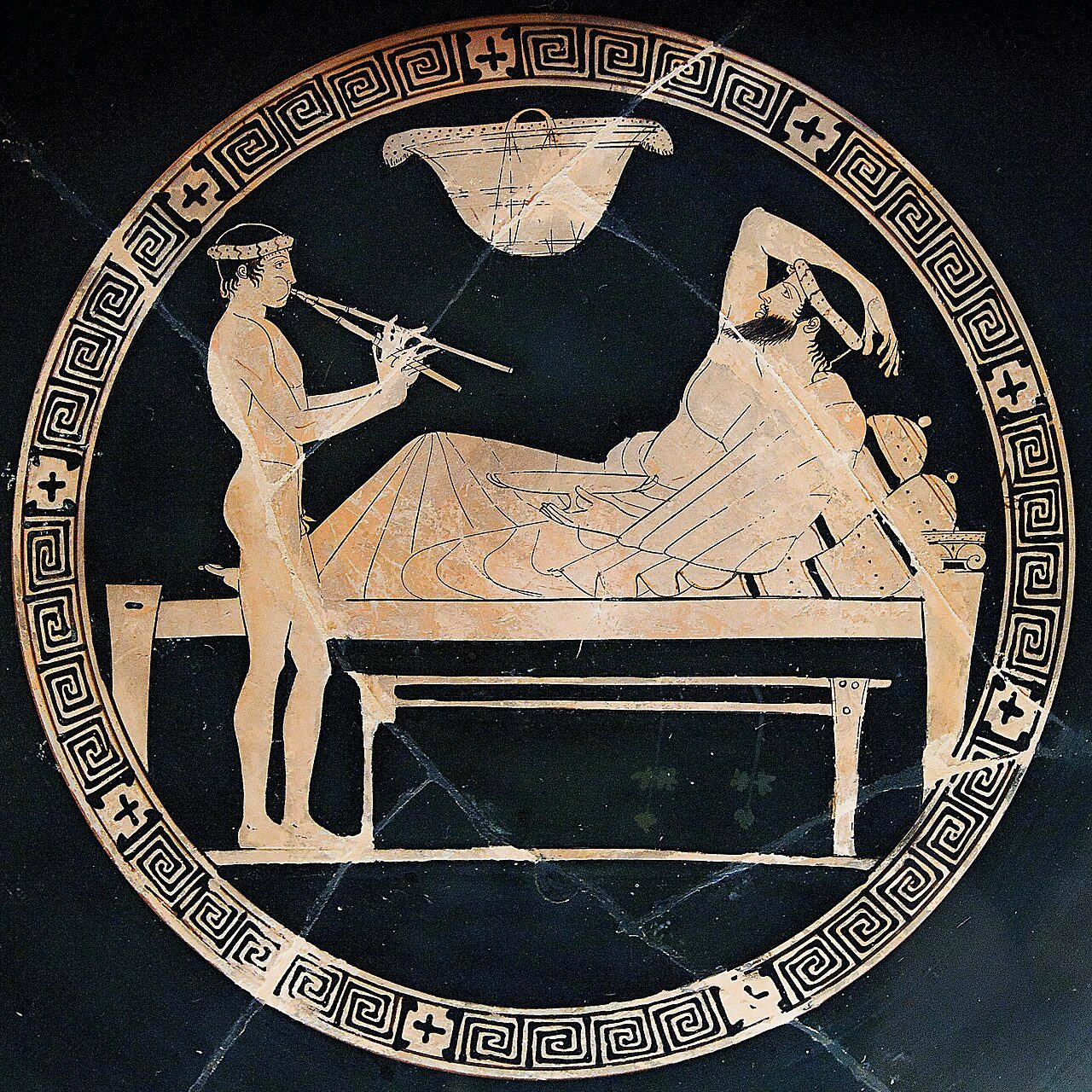 Banquet scene: man reclining on a bench and youth playing the aulos. Tondo of an Attic red-figure cup.
