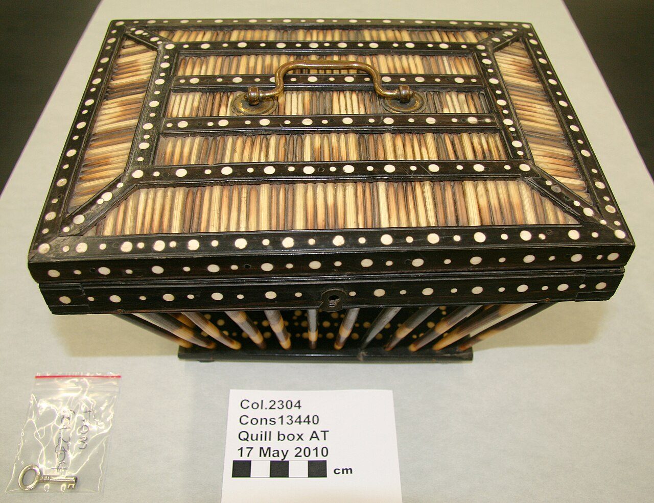 Quill box. Made from porcupine quills and ebony inlaid with ivory discs. Open-sided with a hinged lid. Brass handle at top of lid. Metal lock at front of lid. Underside of lid is lined with blue baize cloth and there is a central decoration of inlaid ivory discs in an ebony…