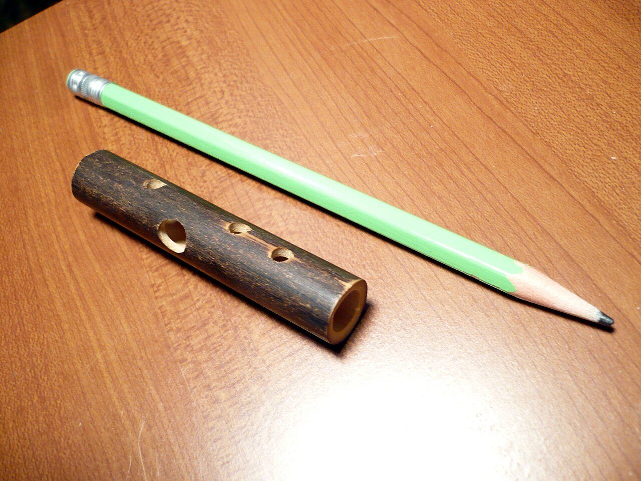 A koudi , with pencil for size comparison. The koudi was probably made in China. The large hole is the blowing hole, and three finger holes are also seen. The holes on both ends of the tube are also used, played with the thumbs.